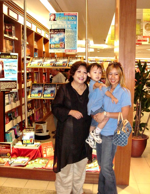 Lillian Too, with daughter Jennifer and grandson Jack, posing under and next to posters of her book, “Get Rich With Water”.