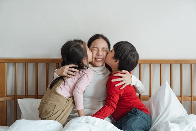 Two children are kissing their mother on the bed
