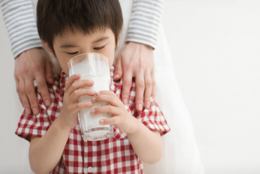 A cute Asian toddler is drinking a big glass of organic milk.