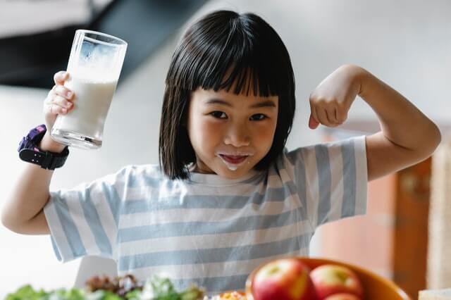 An asian girl lifts her arm with a glass of milk in hand.