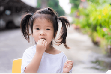 A 15-month-old Asian toddler is eating fruits in front of her house.