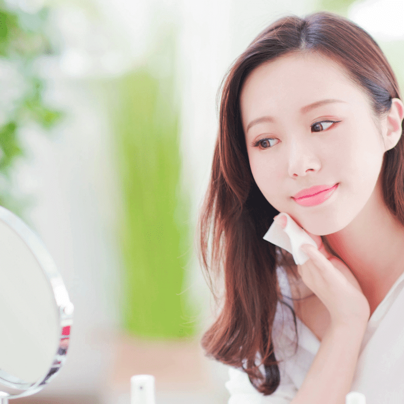 An Asian woman is applying moisturizer in front of the mirror.