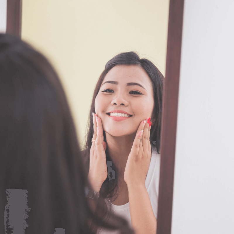 An Asian woman is applying sunscreen on her face in front of the mirror.