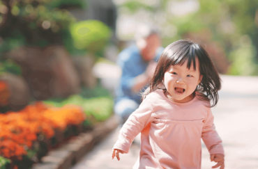 An Asian little girl is running happily with her grandfather sitting behind.