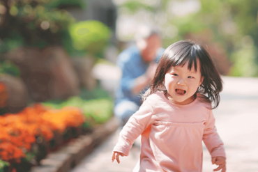 An Asian little girl is running happily with her grandfather sitting behind.