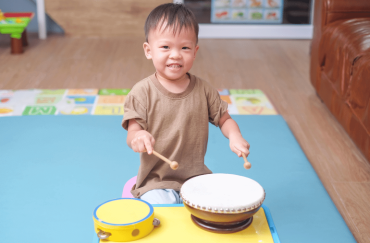 A cute smiling Asian toddler is holding a pair of sticks to play the drums.