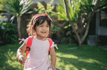 An Asian toddler is running happily in the garden with her bag in the garden.