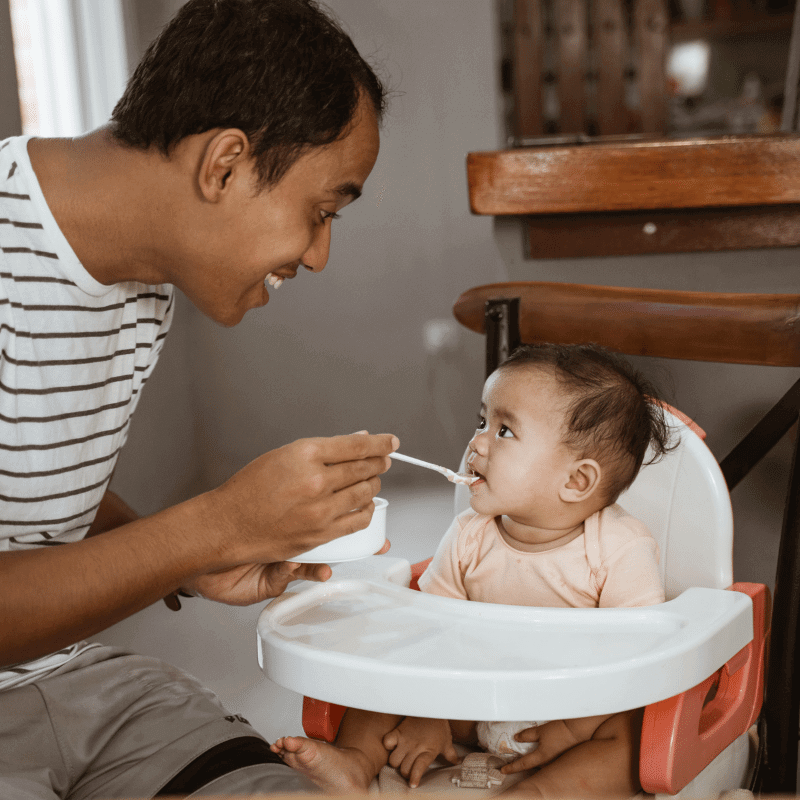 An Asian father is feeding his baby in the high chair.