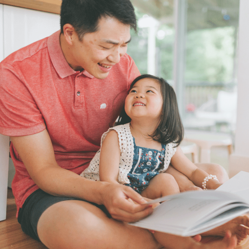 An Asian father is reading a story book to his daughter at home.