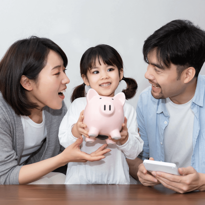 An asian family is smiling with her daughter holding a piggy bank.