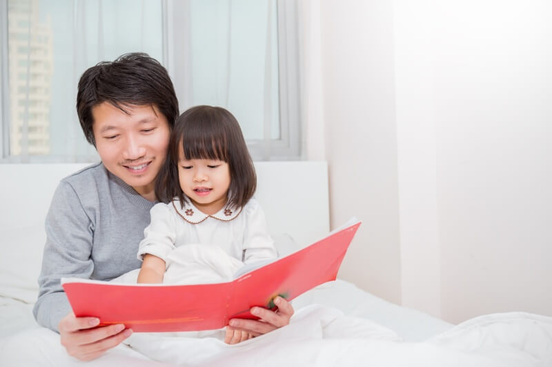 asian father holding story book teaching daughter little girl in bedroom