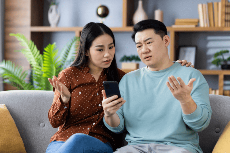  couple sitting on couch looking at phone with confused expressions, discussing something important.
