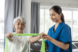 Young physical therapist caregiver assisting mature asian woman grey hair doing exercise with elastic bands at physiotherapy clinic.