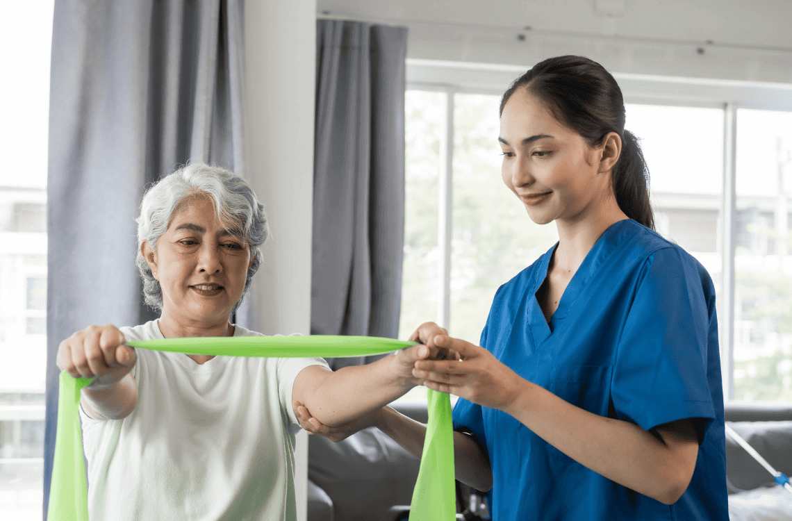 Young physical therapist caregiver assisting mature asian woman grey hair doing exercise with elastic bands at physiotherapy clinic.