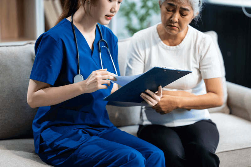 Young Asian nurse attends to older grey-haired mature woman sitting on sofa, conducting health assessment and discussing conditions osteoporosis,