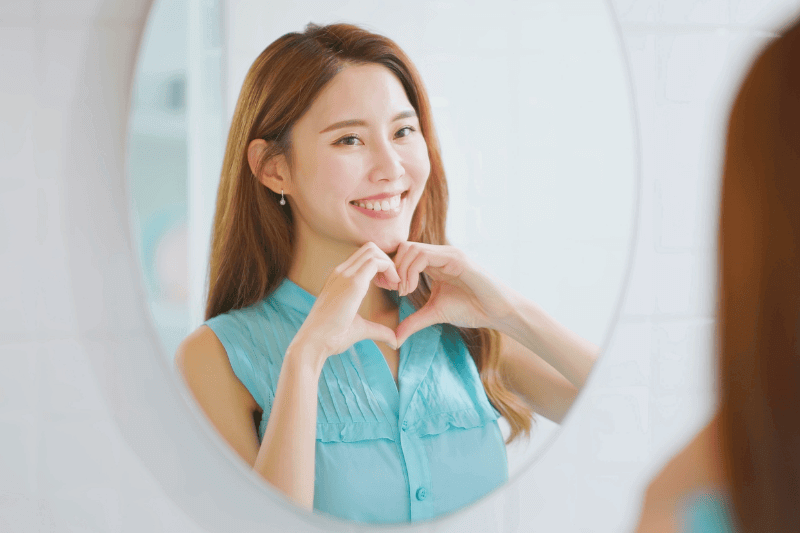 asian cheerful smile woman is gesturing a heart shape with hands while looking in mirror