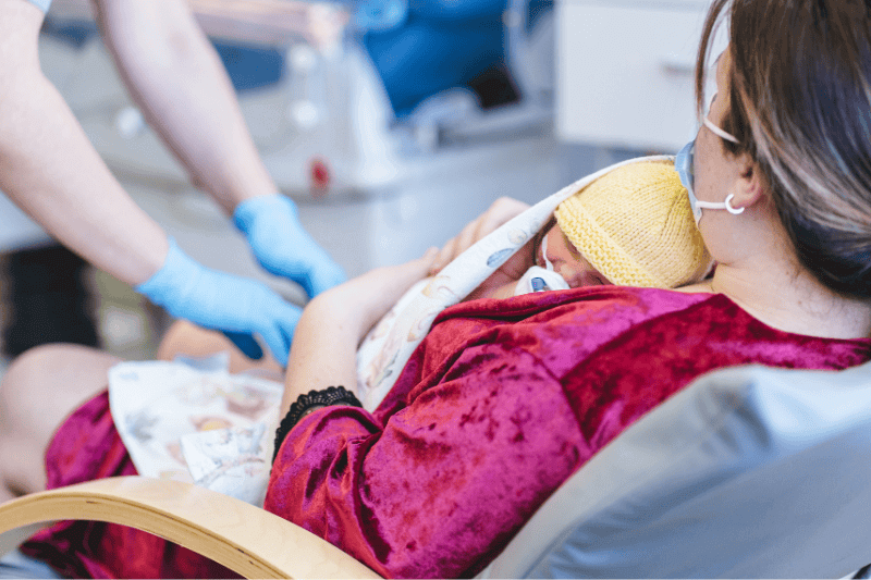 A neonatal nurse places an oxygen mask on a premature baby held by the mother in a neonatal intensive care unit. The baby is wrapped in a blanket and wearing a knitted hat.