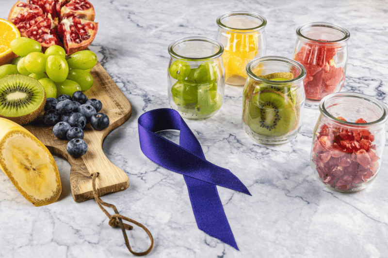 Fruit board and fruit jars with kiwi, pomegranate, grape, orange, grapefruit, blueberry and banana. Superfood against diseases and cancer. A purple ribbon. Concept of fight against cancer