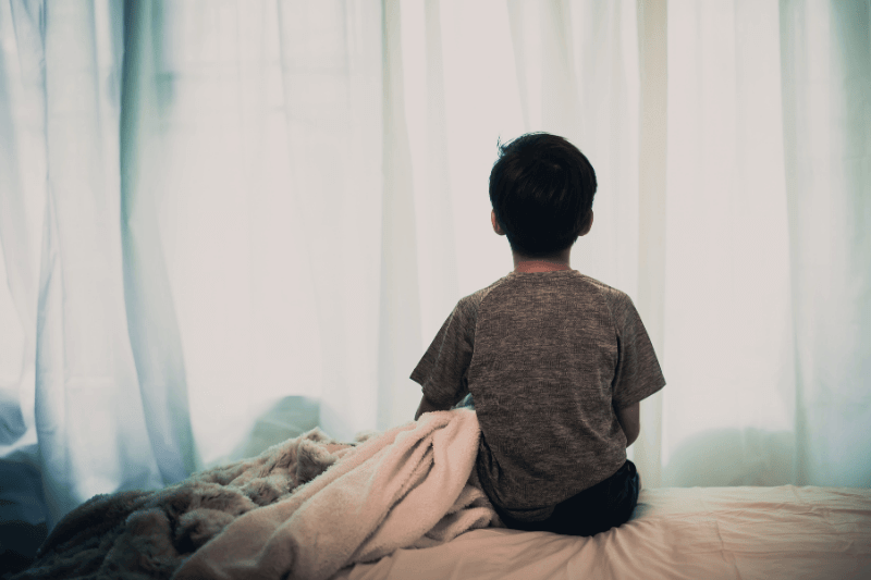 Dark tone color and sad mood of young Asian Thai boy sitting alone on a white bed in a white bedroom