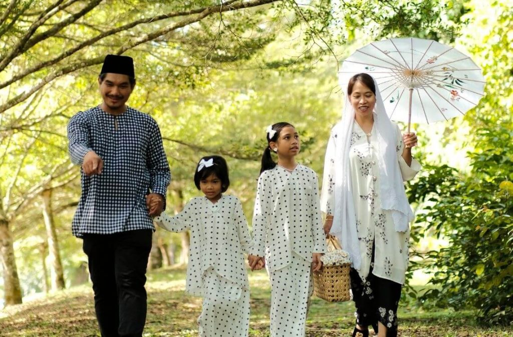 Happy Malay family dressed in traditional attire walking together in a green park, celebrating Hari Raya.