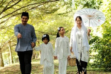 Happy Malay family dressed in traditional attire walking together in a green park, celebrating Hari Raya.