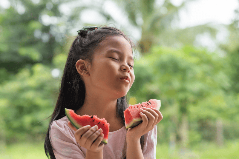 girl with tan skin and pink dress, close the eyes, hold 2 hands the bite to 2 pieces of watermelon