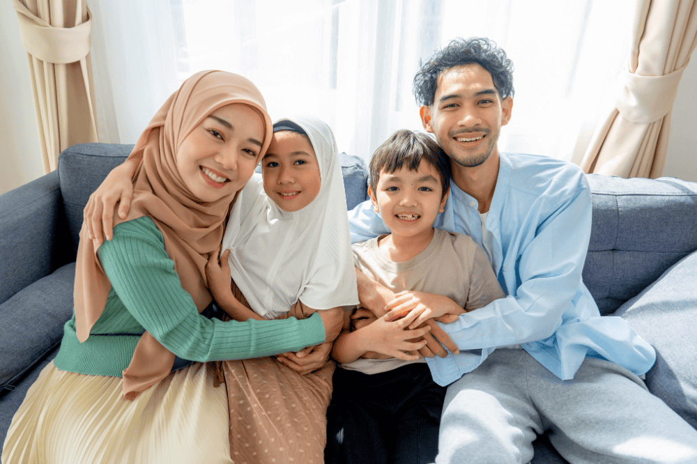 Asian muslim family compose of young mother, father, daughter and son hug and stay together on sofa in living room of their house and look at camera with smiling.