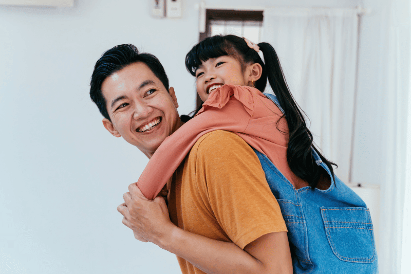 Young father giving his daughter piggyback and smiling in front of house.