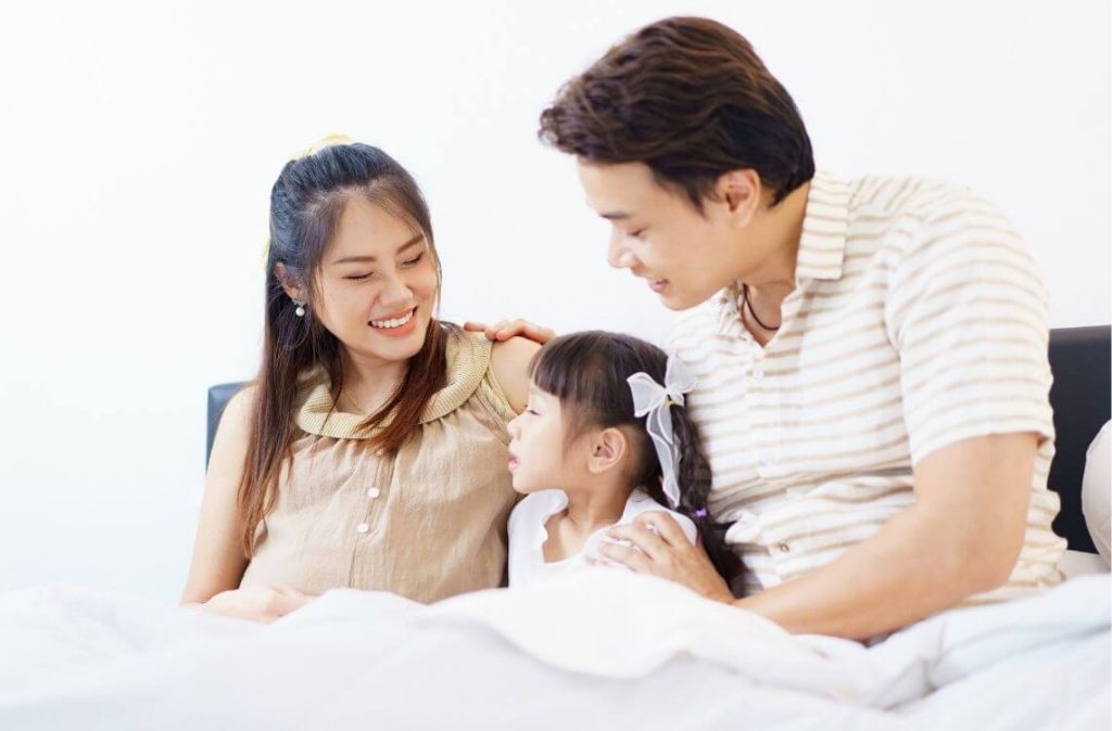 Happy Asian family with father, pregnant mother, and daughter are relaxing or lying down on the bed and talking together. A concept of happiness in family.