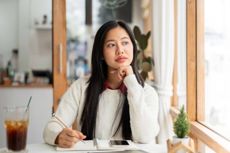 An asian woman is thinking while writing with a pencil in notebook with a smartphone and a tablet place on the table. Study or doing an assignment at cafe or coffee shop.