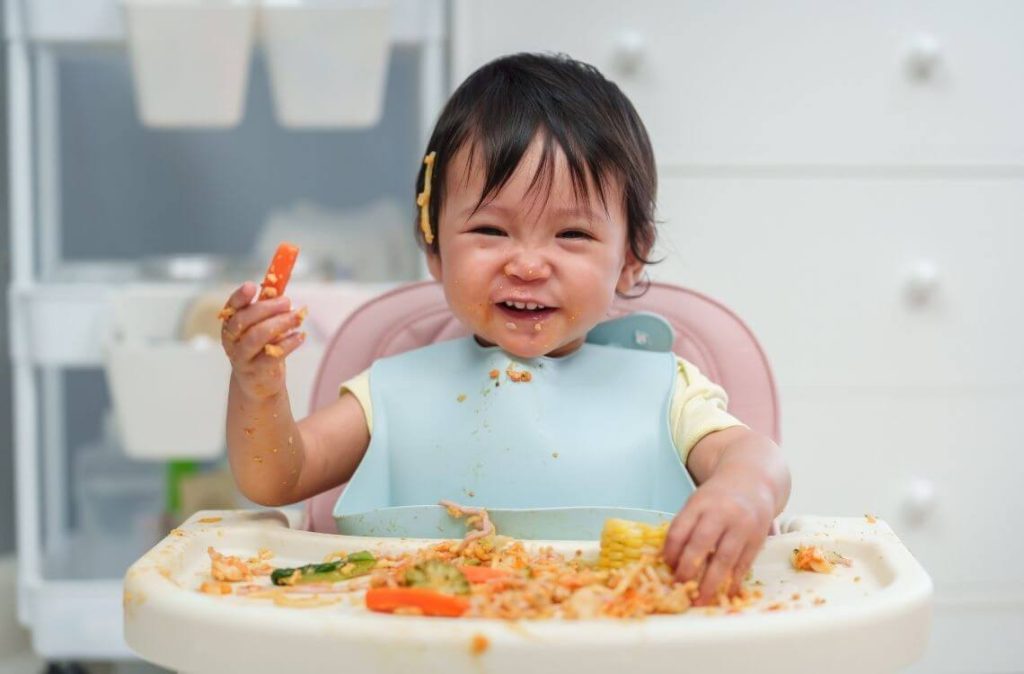 happy infant baby eating food and vegetable by self feeding BLW or baby led weaning on a chair