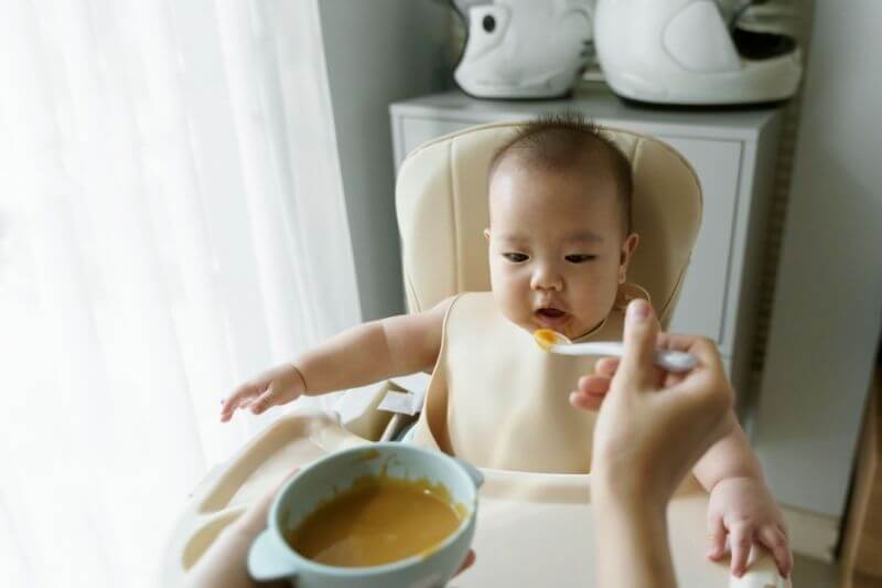 Asian Mother feeding her little baby boy with a spoon. Mom preparing healthy pureed pumpkin for first food in the bowl.