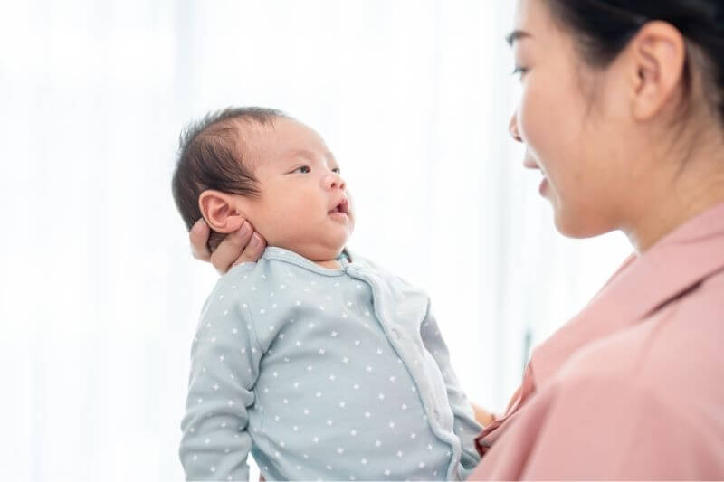 A happy young Asian mom is talking and comforting her infant baby son while holding him in her arms, standing by the window in the living room. mother's love and tenderness, motherhood