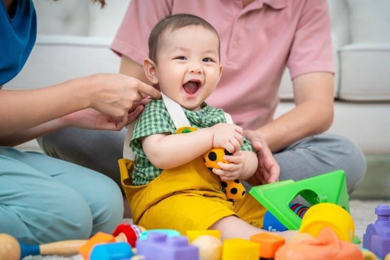 A family of three, a man and two women, are playing with toys on the floor. The baby is holding a toy truck and the parents are clapping. Scene is happy and playful