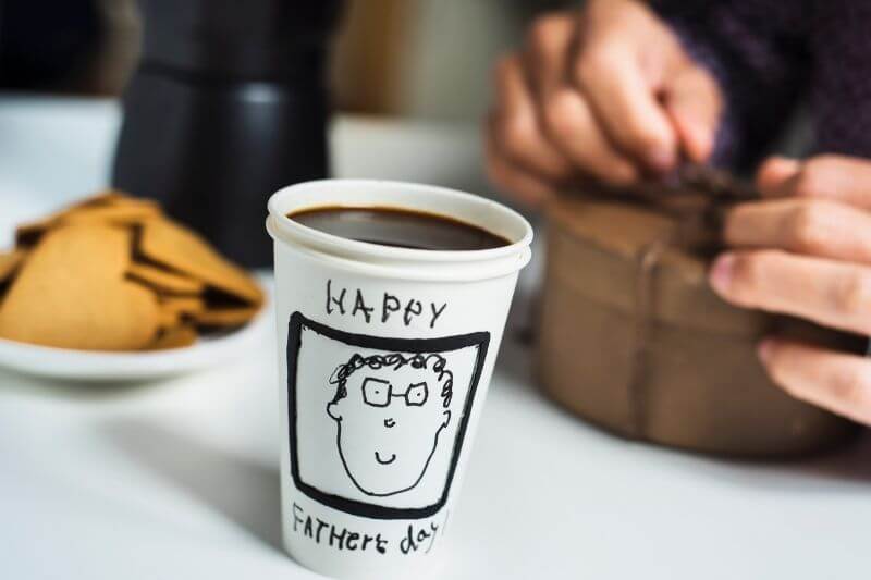 closeup of a disposable cup with coffee, with the text happy fathers day handwritten in it, on a table set for breakfast and a caucasian man opening a gift in the background