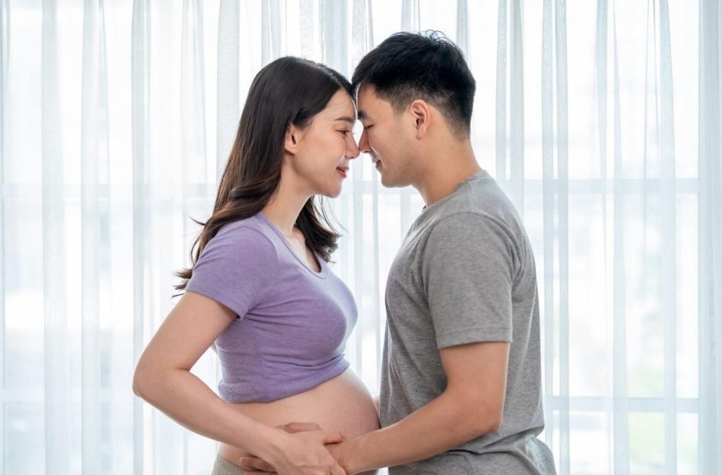 Asian young pregnant woman gently stroke belly with husband in house. Attractive expectant couple touch baby bump and share loving moment, ready to become parents in future in living room at home.
