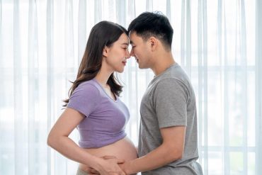 Asian young pregnant woman gently stroke belly with husband in house. Attractive expectant couple touch baby bump and share loving moment, ready to become parents in future in living room at home.