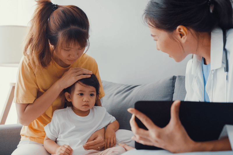 Young Asia female pediatrician doctor and little girl patient using digital tablet sharing good health test news with happy mom sit on couch in house. Medical insurance, Visit patient at home concept.