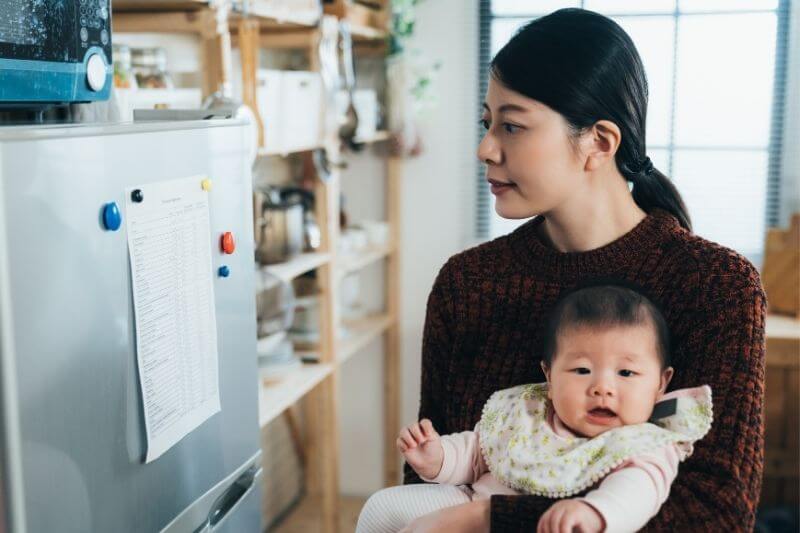 asian cute infant girl is looking at the camera with innocence in her mother’s arm while her mother is checking a list on refrigerator in the kitchen at home.