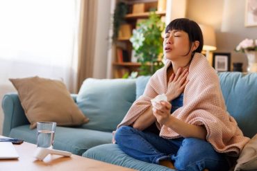 Middle-aged Asian woman relaxing at home while sick, sitting on the couch covered with blanket having fever and sore throat