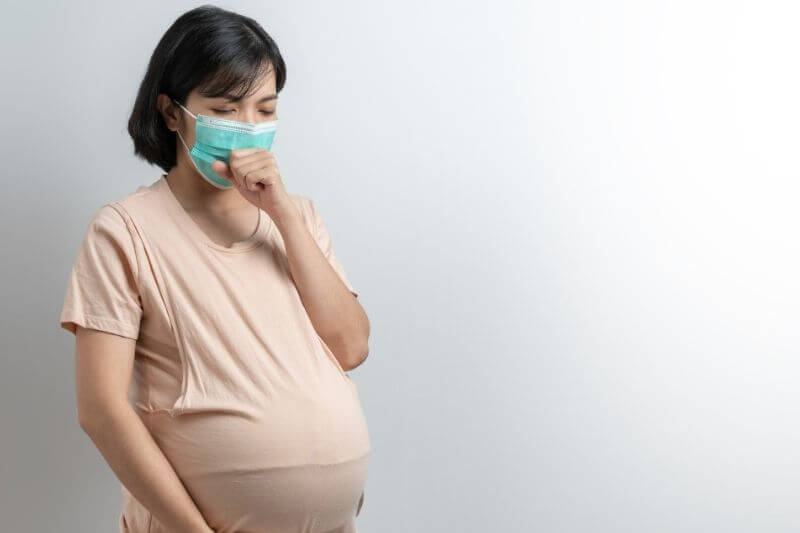 Portrait of Asian Pregnant woman wearing protective face mask coughing on white isolated background. Young mother having a cold or flu standing touching her belly with copy space during covid pandemic