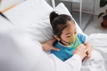 blurred doctor adjusting oxygen mask on sick asian child on hospital bed