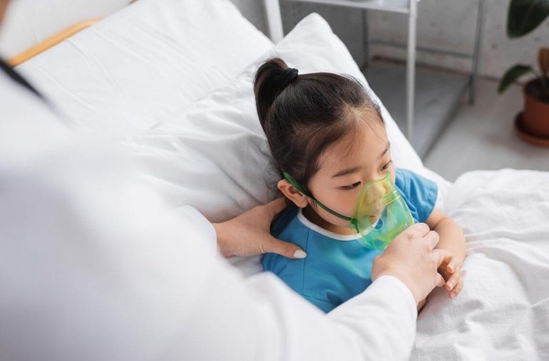blurred doctor adjusting oxygen mask on sick asian child on hospital bed