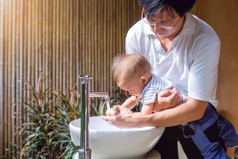 Father washing / cleaning Cute little Asian toddler baby boy child hands on white sink and water drop from faucet, Selective focus at hand with running water from faucet, Sanitation / hygiene concept