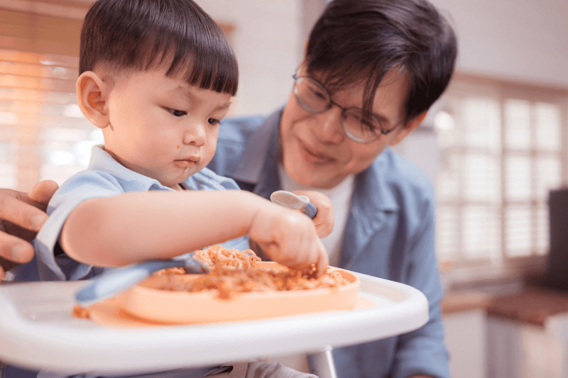 Adult supervising a toddler eating while seated upright in a high chair.